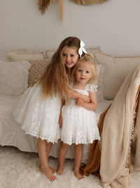 A big sister hugs her little sister, they are wearing matching flower girl outfits, our Loulou lace flower girl dress and Loulou baby flower girl romper.