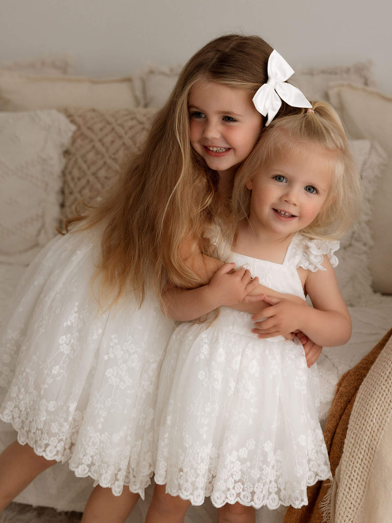 An older sister hugs her toddler sister, they are wearing matching white flower girl dresses. The older sister wears our Loulou flower girl dress and the younger sister wears our Loulou flower girl romper.