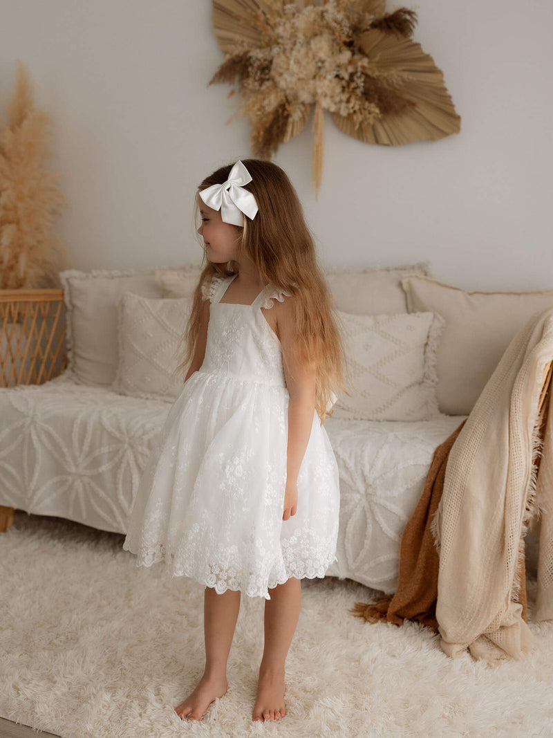 A little girl stands smiling, she is wearing an ivory lace flower girl dress, our Loulou dress, and an ivory satin bow hair clip.