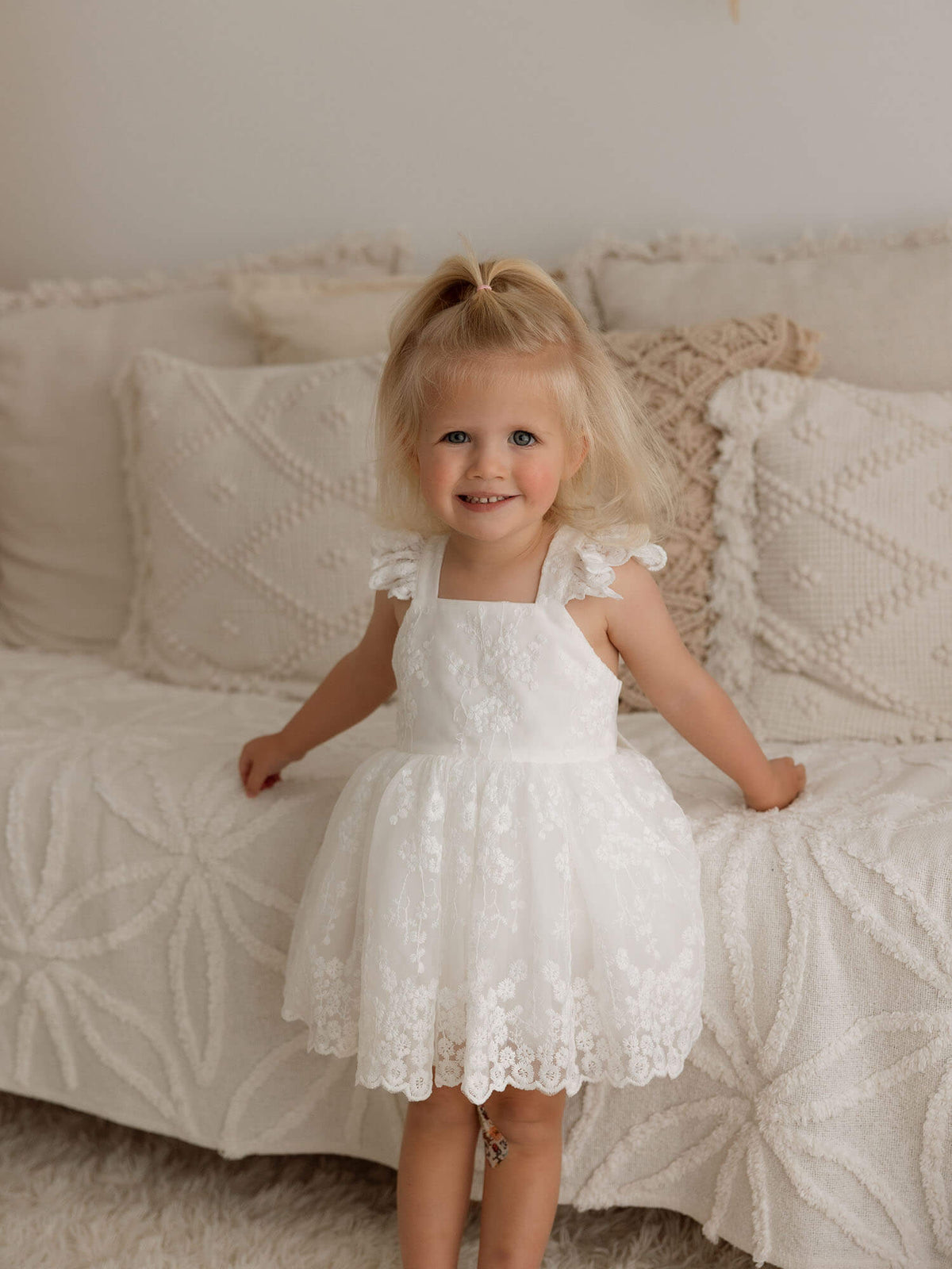 A toddler stands wearing our ivory lace baby flower girl dress, Loulou.