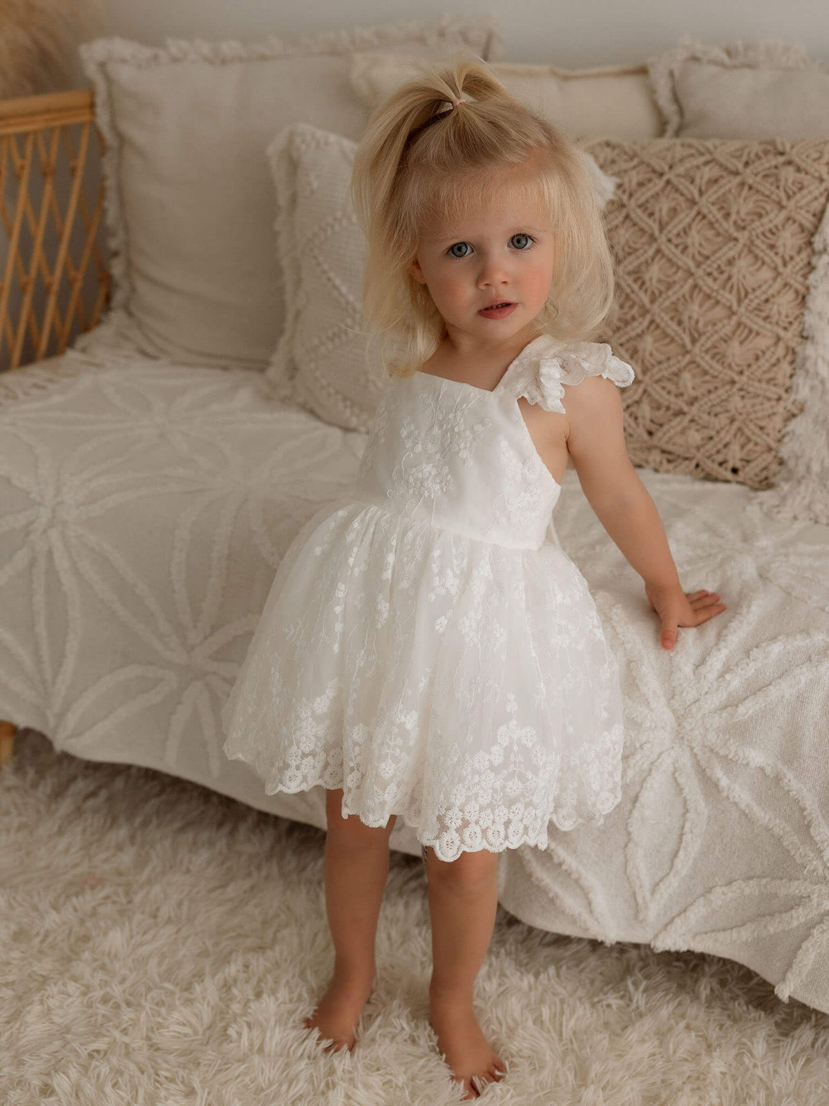 A toddler wears our ivory Lace Loulou flower girl romper with lace flutter sleeves.