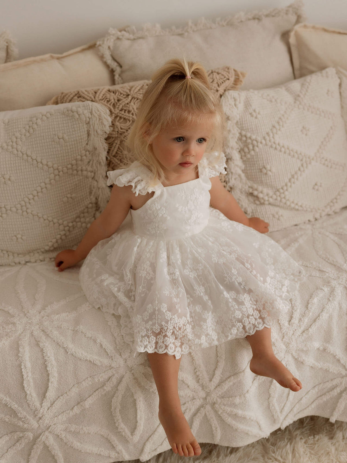 A toddler sits wearing our lace Loulou baby flower girl dress, showing the lace detail on the dress.