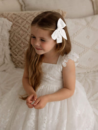 A little girl sits wearing our Loulou natural white lace flower girl dresses and ivory satin bow hair clip.