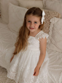 A little girl sits wearing a white flower girl dress, our Loulou dress, along with a satin bow in her hair.