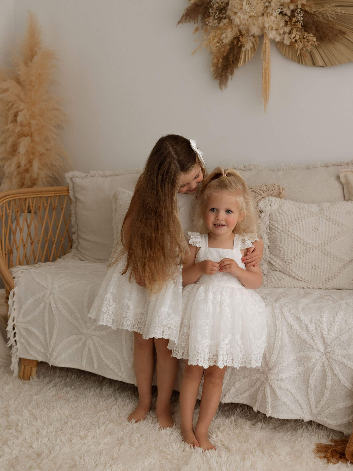 An older sister wearing our Loulou flower girl dress, hugs her little sister who is wearing our matching Loulou flower girl romper for toddlers.