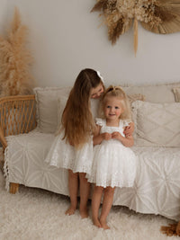 An older sister wearing our Loulou flower girl dress, hugs her little sister who is wearing our matching Loulou flower girl romper for toddlers.