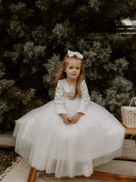 A young flower girl sits on a bench outside wearing our Lucy flower girl dress,showing the full tulle skirt and full length satin sleeves.