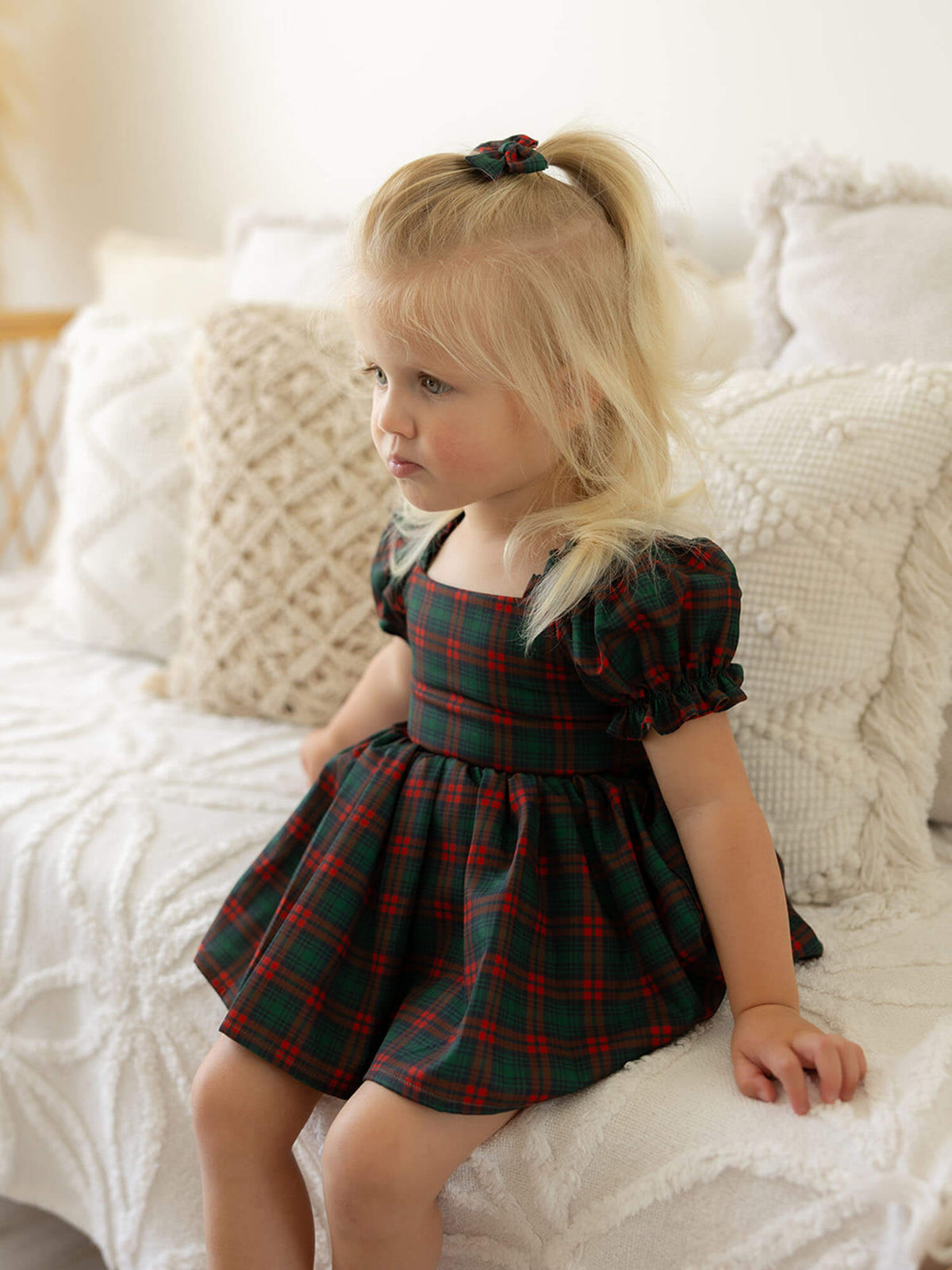 A toddler sits wearing our Mila tartan Christmas romper and matching bow clip in her hair.