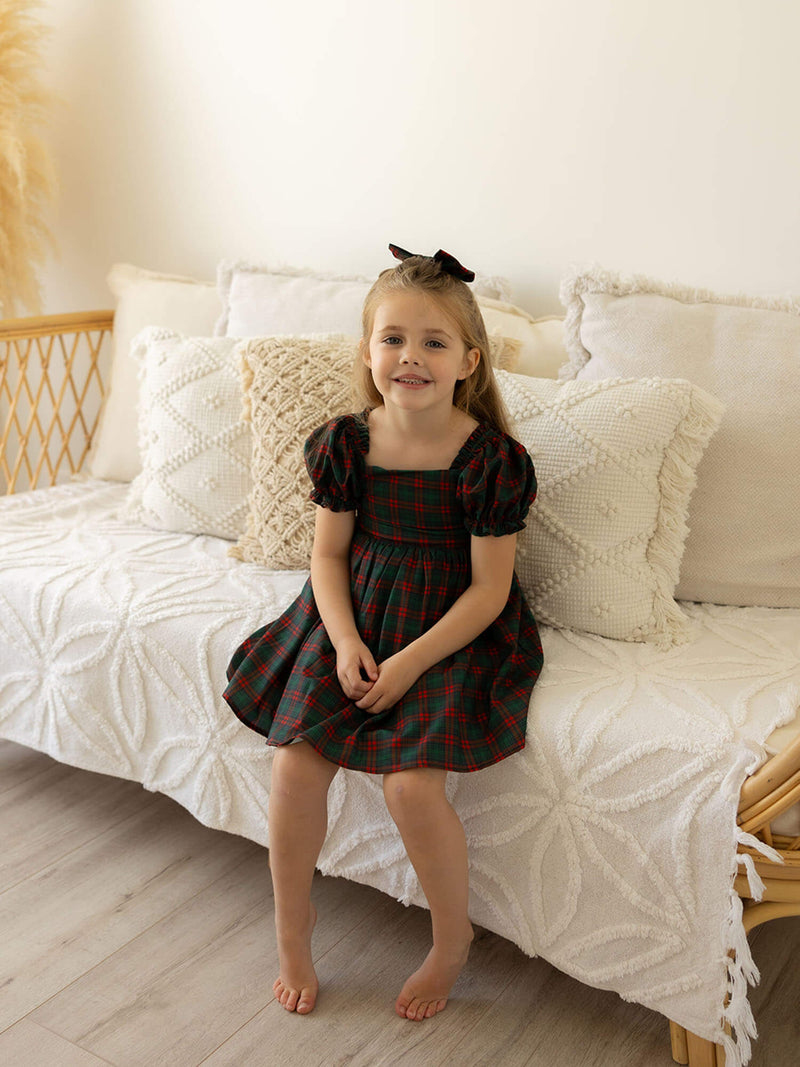 A young girl sits wearing our Mila girls tartan Christmas dress and matching tartan bow in her hair.