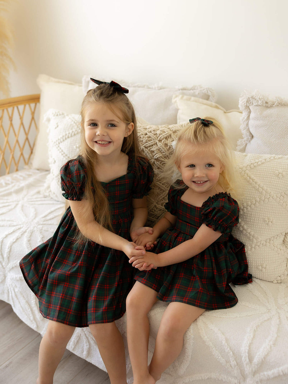 Sisters sit holding hands, one wears our Mila girls Christmas dress and the other wears our Mila Christmas baby romper.