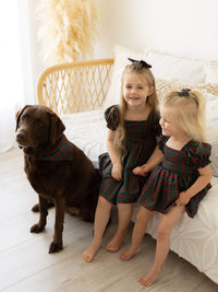 Two sisters and a dog sit wearing matching Christmas dresses and accessories in our Christmas plaid print.