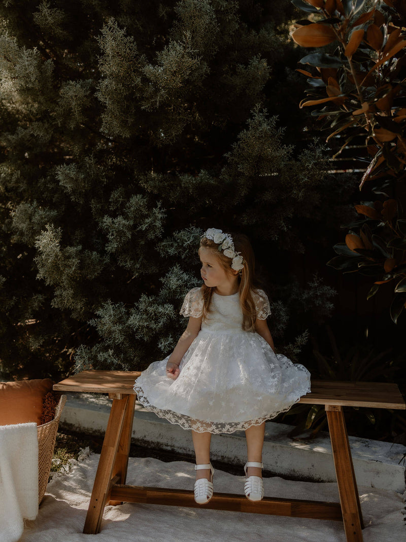 Pippa flower girl dress in floral lace is worn by a young girl sitting on an outdoor seat.
