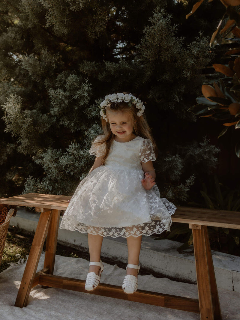 Pippa ivory floral lace flower girl dress is worn by a young girl, she sits on a stool laughing, wearing a rose flower crown in her hair.