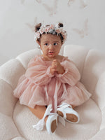 Wren baby flower girl dress in dusty pink is worn by a toddler who is sitting on a seat. She wears a matching flower crown, our Rose blush flower crown.