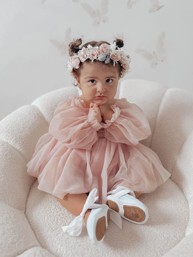 Toddler sits wearing our dusty pink baby flower girl romper, Wren romper, with her hands at her chin showing the billowing soft tulle sleeves. She also wears our Rose flower crown in blush. 