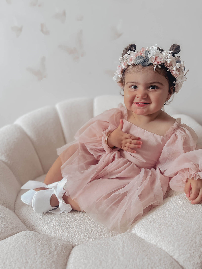 Wren baby flower girl romper with full length tulle sleeves is worn by a smiling toddler. She also wears our Blush Rose flower crown with pigtails in her hair.