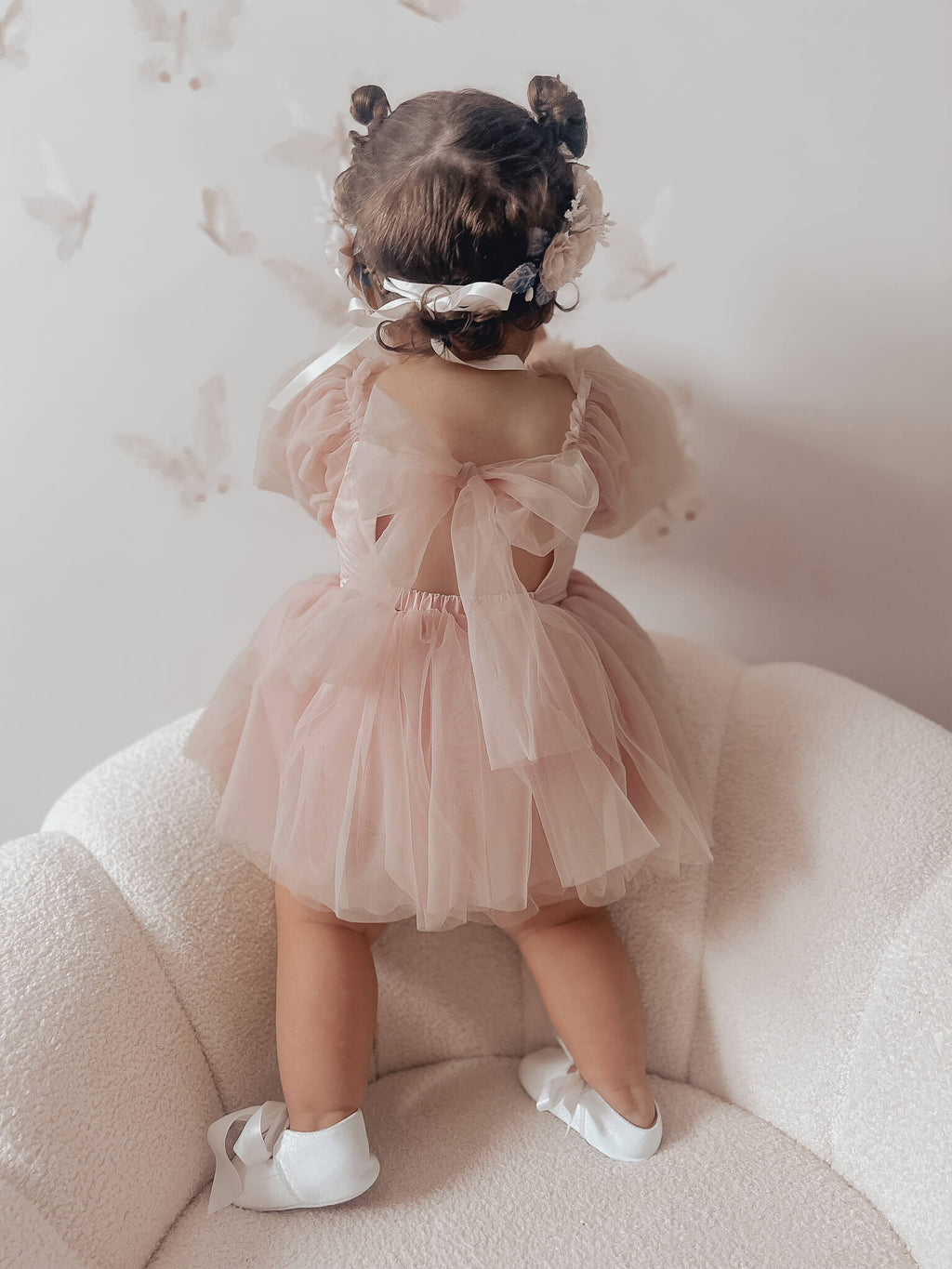 Toddler sits wearing our dusty pink baby flower girl romper, Wren romper, with her hands at her chin showing the billowing soft tulle sleeves. She also wears our Rose flower crown in blush. 