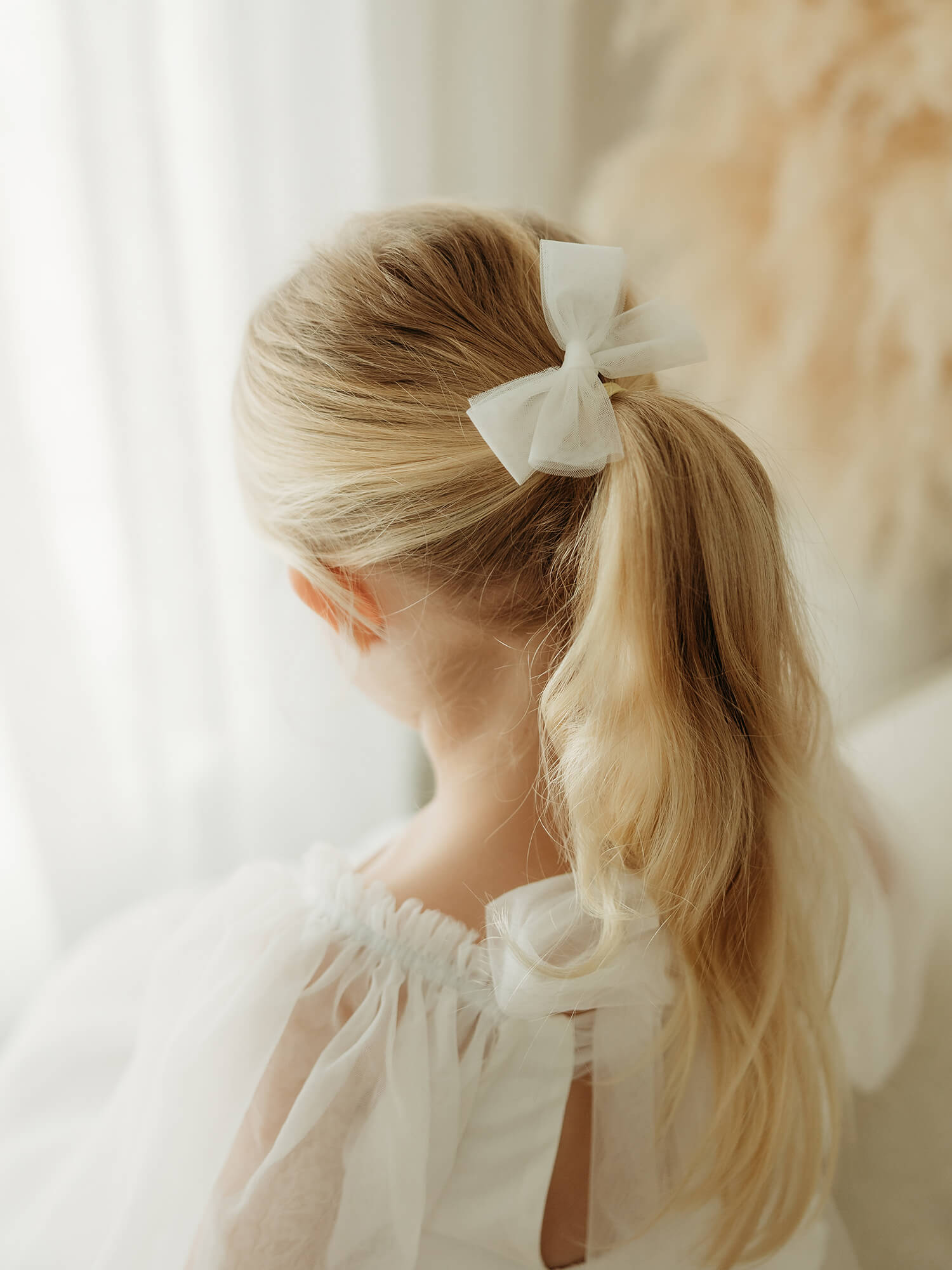 Child with blonde hair in a ponytail with a white tulle bow, wearing a white tulle flower girl dress.