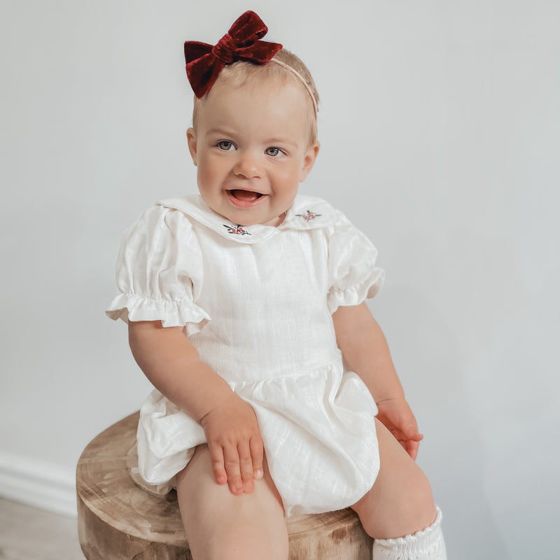 Emma puff sleeve Christmas romper is worn by a smiling baby, who also wears a velvet red bow hedband.