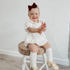 Emma linen Christmas romper is worn by a smiling baby sitting on a stool.