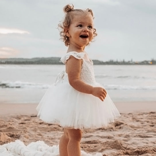 Young girl wears our Everly lace baby romper with tulle skirt in ivory. She stands on the beach.