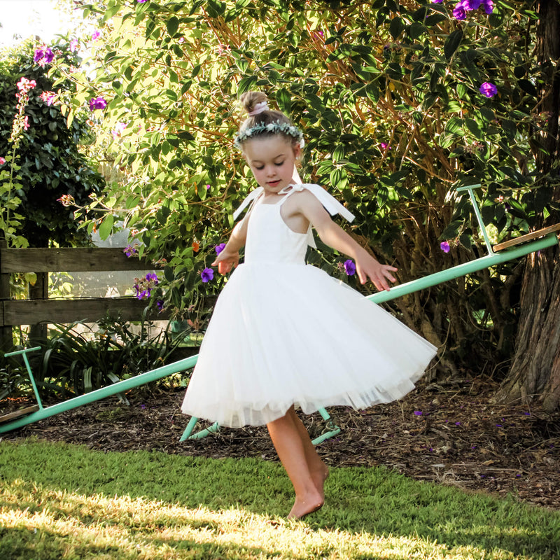 Margot ivory flower girl dress on a young girl who twirls around outside. It has a tea length tulle skirt, and tie straps. She also wears our Eden flower crown.