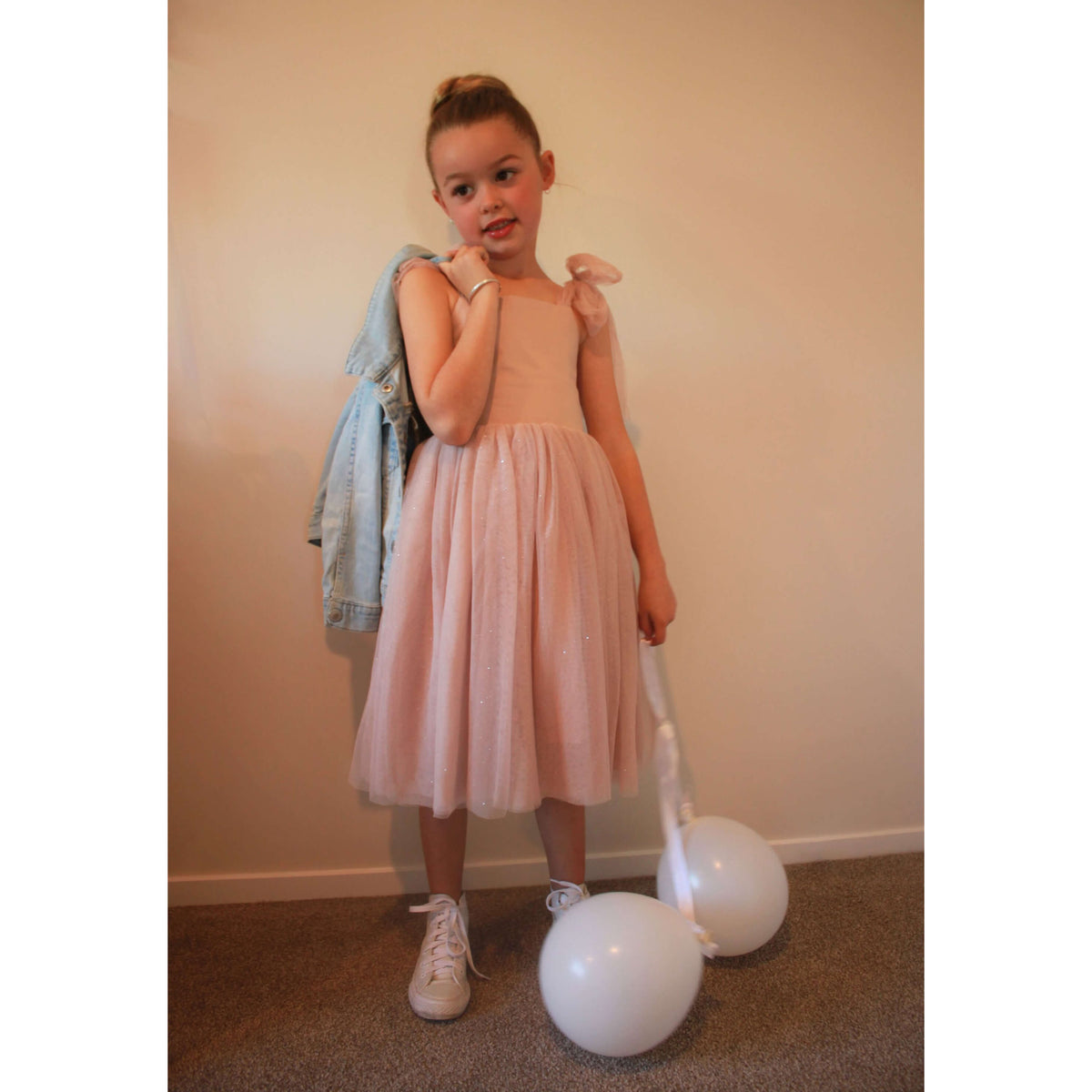 Rosie tulle girls dress worn by a young girl. She holds a denim jacket over her shoulder with balloons at her feet. The tulle skirt of this flower girl dress has some glitter in it.