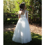Sonnet ivory flower girl dress shown from the back on a young girl. She stands in a garden, wearing the full length ivory flower girl dress and a flower crown. The back of the dress is shirred and has bow tie straps.