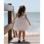 Briar lace flower girl dress shown from the back on a young flower girl, at the beach. Showing the full length lace sleeves, with scoop back and knee length tulle skirt.