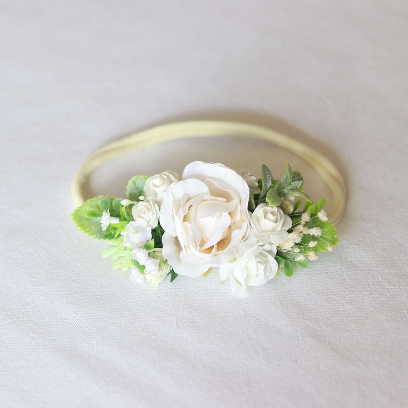 Delilah girls floral headband with a central ivory flower, surrounded by smaller ivory flowers and greenery, on a soft elastic headband.