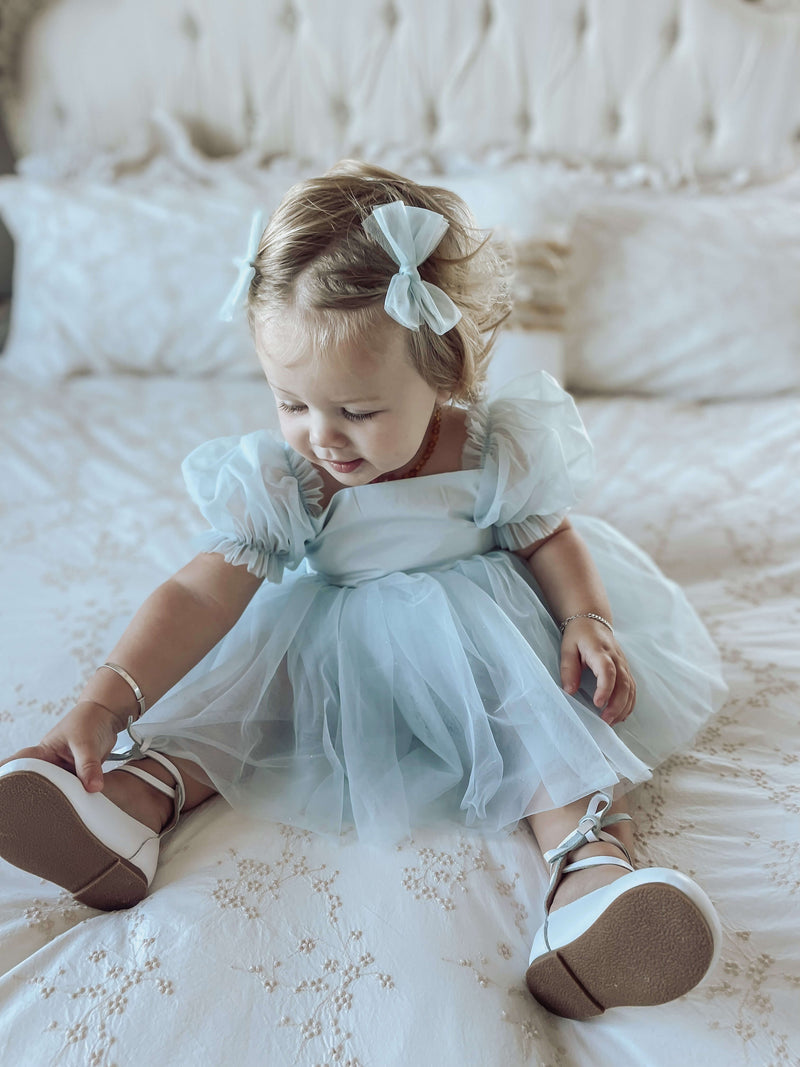 Gabrielle baby flower girl dress is worn by a toddler, in colour dusty blue, along with matching tulle pigtail bows in her hair.