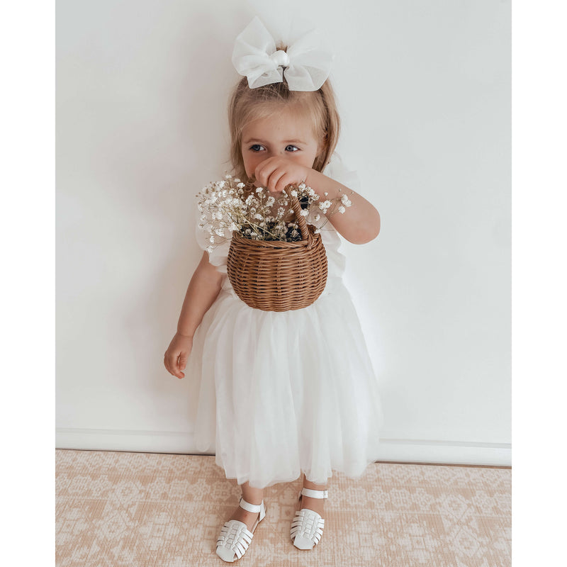 A young flower girl holds a basket of flowers while wearing our Gigi flower girl dress in ivory, and an ivory tulle bow in her hair.