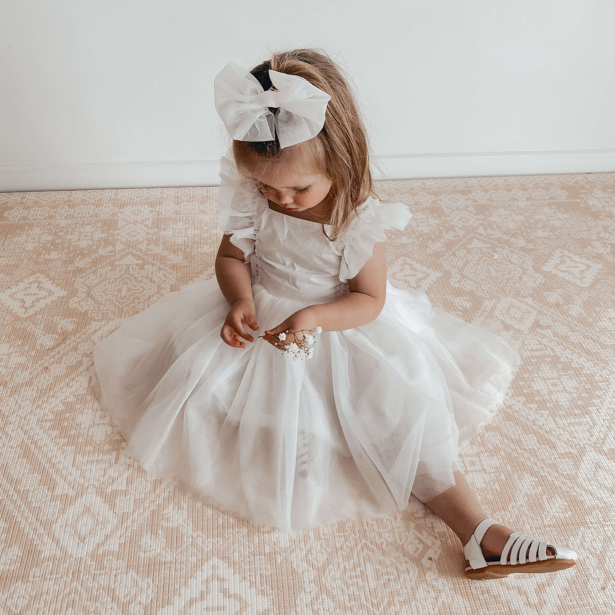 Gigi flower girl dress and tulle bow is worn by a young girl who sits on the floor holding some flowers.