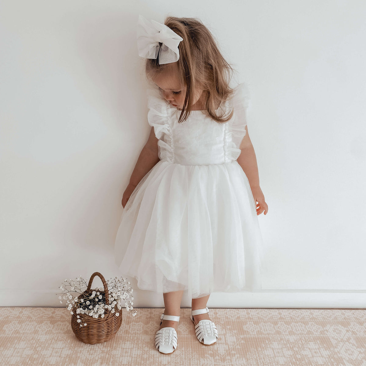 Gigi white flower girl dress worn by a young girl, she also wears our ivory tulle bow in her hair and stands beside a basket of flowers.