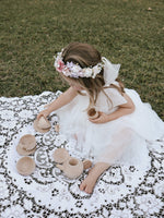 Harper ivory flower girl dress in tea length is worn by a young girl who is having a tea party outside. She sits on a lace blanket and wears our Luna flower crown in her hair.