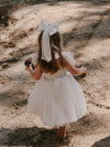 A young girl wears our large classic tulle bow in ivory and our ivory Harper flower girl dress in tea length, shown from behind. The flower girl dress has shirring at the back for stretch.
