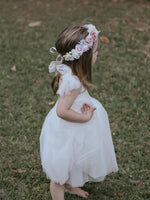 Harper tea length flower girl dress is worn by a young girl, she also wears our girls Luna flower crown. The dress is white and has tulle tie straps, which are tied in bows at her shoulders.