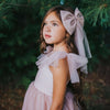 Rosie dusty pink flower girl dress shown close up being worn by a young girl, the tulle straps are tied in bows. She wears a large dusty pink tulle bow in her hair.