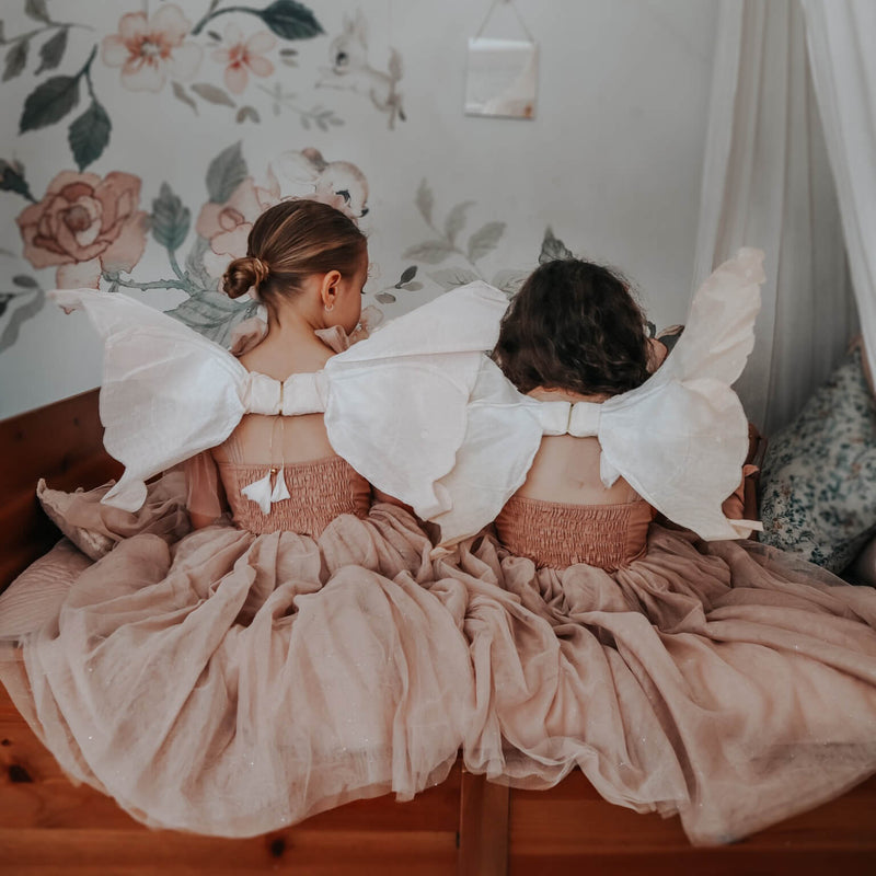 Sisters sit wearing fairy wings and our Rosie tulle dress, shown from the back. Showing the champagne tulle skirt and shirred back.