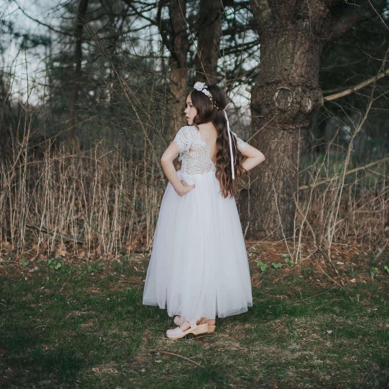 Sophia lace flower girl dress in blush being worn by a young girl. She stands outside, wearing the full length tulle dress with lace bodice, and a blush flower crown. The flower crown is our Amelie flower crown in blush which matches our Sophia blush flower girl dress perfectly.